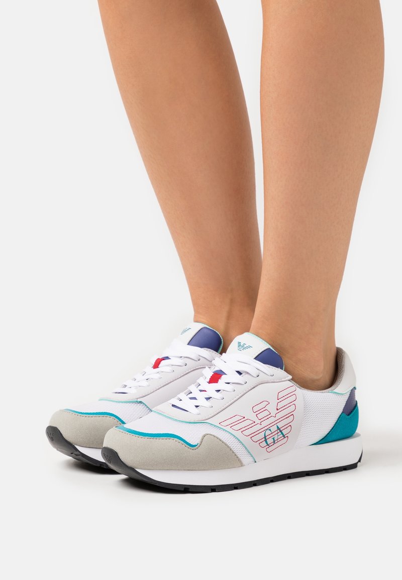 Scarpe da ginnastica bianche con accenti turchesi e grigi, realizzate in materiali mesh e scamosciati, con cuciture a contrasto e un design del logo a fantasia.