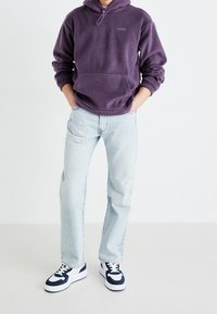 Persona con felpa con cappuccio in pile Levi's viola, jeans azzurri e sneakers bianche con dettagli blu navy, in piedi con le mani in tasca su sfondo bianco.