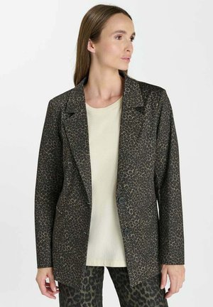 Vrouw draagt een donker leopardprint blazer over een beige blouse en bijpassende leopardprint broek, kijkt opzij tegen een effen achtergrond.