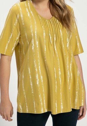 Femme portant un haut jaune moutarde à manches courtes avec des motifs à rayures verticales blanches et des plis près de l'encolure ronde.