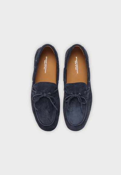 Paire de mocassins en daim bleu foncé avec nœuds sur le devant et semelles intérieures beige portant l'étiquette "Stuart Weitzman Made in Italy."