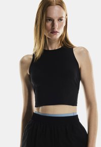 Top de tank noir court avec un col haut et sans manches ; fabriqué dans un matériau lisse et extensible. Assorti à un bas noir ample.