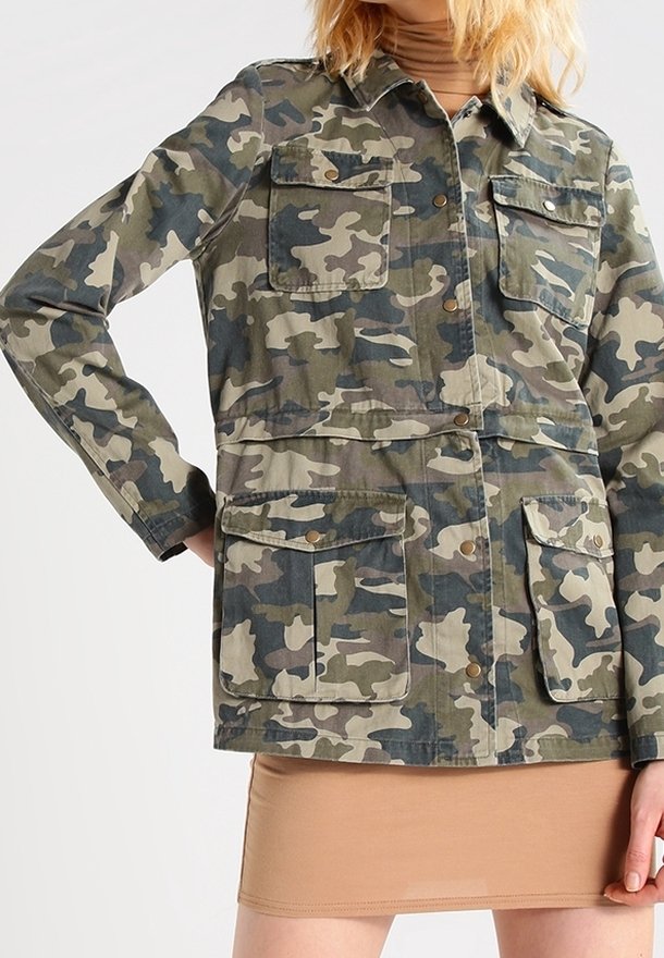 Woman wearing green and brown camouflage jacket with buttoned flap pockets over a beige skirt, hand on hip.