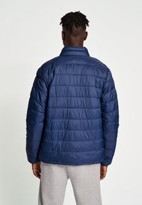 Mörkblå puffjacka med kviltad textur, hög krage och en avslappnad passform. Har horisontella sömmar och en slät syntetisk yta.