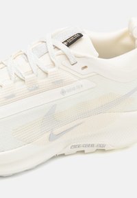 Baskets blanches Nike avec un dessus texturé, étiquette GORE-TEX, accents en maille et un logo visible en gris. Le design présente une silhouette massive.