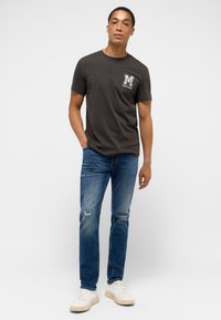 Mörkgrå crew neck t-shirt med vit "MUSTANG" logotyp på bröstet, kombinerad med ljus tvätt slim-fit jeans med slitna detaljer vid knäna och vita sneakers.