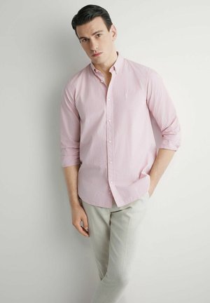 Mężczyzna w jasnoróżowej, kratkowanej koszuli na guziki z podwiniętymi rękawami i jasnobeżowych spodniach, opierający się o biały mur.