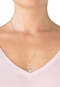 Halskette mit goldfarbener Anhänger in minimalistischer Gesichtsform, aufgehängt an einer zarten Kette, getragen über einem hellen Oberteil.