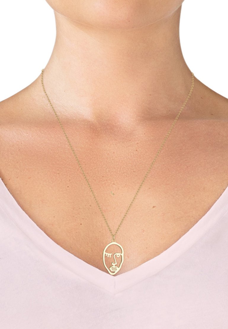 Halskette mit goldfarbener Anhänger in minimalistischer Gesichtsform, aufgehängt an einer zarten Kette, getragen über einem hellen Oberteil.