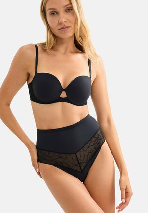 Femme portant un soutien-gorge noir sans bretelles et une culotte noire taille haute en dentelle, debout avec une main sur la hanche devant un fond uni.