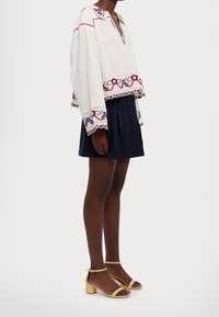 Persona vestida con una blusa blanca de manga larga con bordado rojo y azul, una falda plisada azul marino y sandalias de tacón bloque con tira en el tobillo de color beige, de pie de lado.