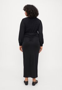 Svart kjole med lange, puffede ermer, teksturert bodice og rynket detaljer ved midjen, som faller til midt på leggen med en rett kant. Sorte sko synlige.