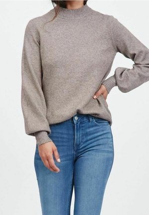 Femme portant un pull en maille taupe à manches longues bouffantes, associé à un jean taille haute bleu, sur un fond blanc.