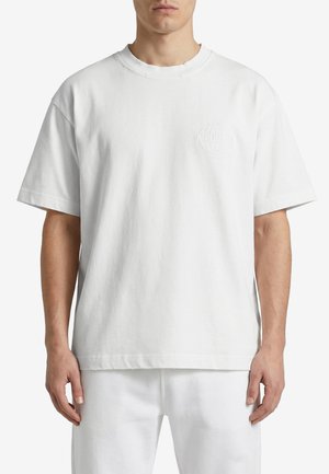 Uomo che indossa una maglietta bianca a maniche corte, larga, con un sottile logo in rilievo sul petto sinistro, abbinata a pantaloni bianchi.