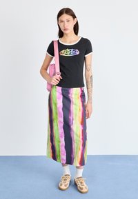 Sort t-shirt med korte ærmer og multicolor logo, parret med en lang nederdel med lodrette striber i pink, grønne og lilla nuancer. NEutrale sneakers og pink taske.