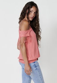 Koralo spalvos off-the-shoulder viršus su nėriniu apdaila ir karoliukų akcentais. Audinys yra tekstūruotas, laisvas įsilaisvinimas, derinamas su šviesiai mėlynomis džinsinėmis kelnėmis.