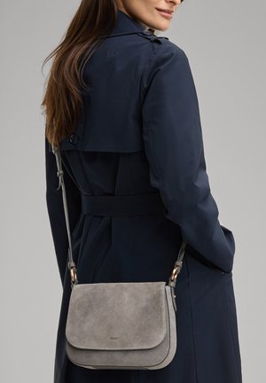 Frau trägt einen marineblauen Trenchcoat mit einer grauen Wildleder-Umhängetasche, die an ihrer Hüfte gegen einen neutralen Hintergrund ruht.