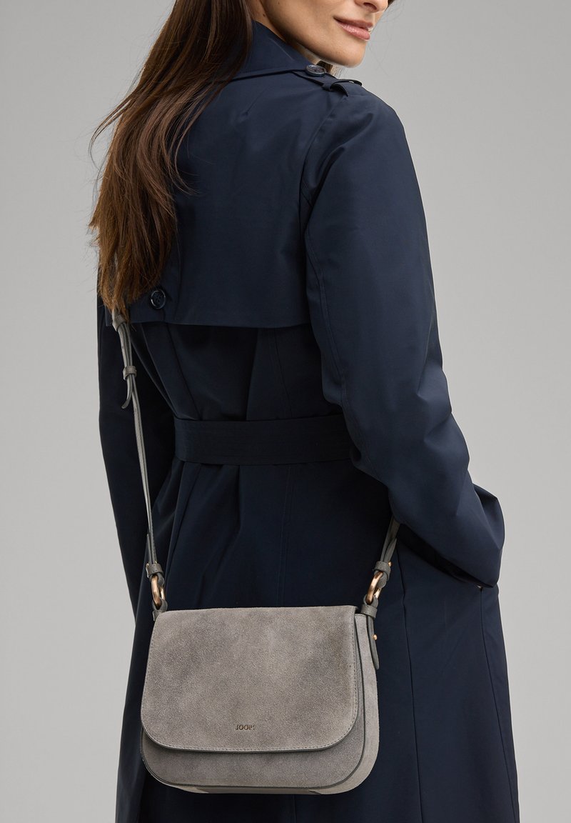 Vrouw die een marineblauwe trenchcoat draagt met een grijze suède schoudertas die op haar heup rust, tegen een neutrale achtergrond.