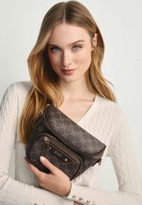 Młoda kobieta z długimi blond włosami, ubrana w białą bluzę, trzyma ciemno-brązową torbę na ramię z wzorem i zamkiem.