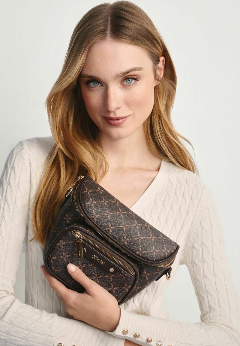 Młoda kobieta z długimi blond włosami, ubrana w białą bluzę, trzyma ciemno-brązową torbę na ramię z wzorem i zamkiem.