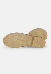 Bota de ante beige con suela de goma texturizada. Punta redondeada y detalles de costura a lo largo de la parte superior. Diseño minimalista con superficies lisas.