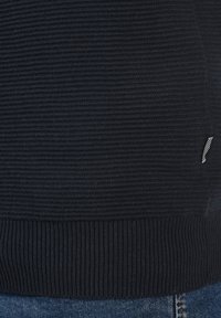 Námořnicky modrý žebrovaný svetr s texturovanou tkaninou a kontrastní bílou proužkovanou visačkou na lemu, podložený denimovým pozadím.