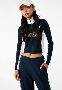 Chaqueta con cremallera recortada en navy y blanco con el texto 'LEGEND'. Tejido suave, mangas largas, combinada con pantalones elásticos navy a la cintura con rayas.
