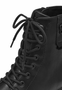 Botín negro de cuero hasta el tobillo con puntera redondeada, ojetes de metal y cordones planos. La superficie tiene una textura suave con detalles de costura mínimos.