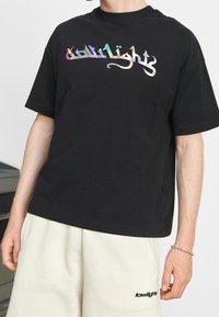 Person in schwarzem T-Shirt mit holografischem "lowlights"-Text und beigen Shorts mit "lowlights"-Logo auf dem linken Oberschenkel, steht drinnen.