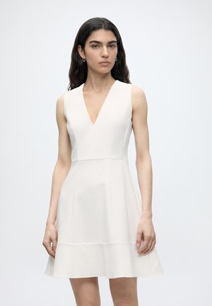 Woman with long dark hair wearing a sleeveless white dress with a V-neckline, standing against a plain light background.