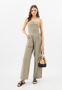 Tutina khaki con design senza spalline, cerniera frontale, pantaloni larghi plissettati, abbinata a una borsa a secchiello bicolore e infradito nere.