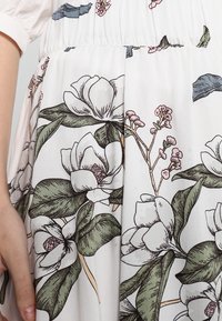 Vitt tyg med ett blommönster som har stora magnoliablommor i dämpad grön och rosa, detaljerade blad och en mjuk, slät yta.