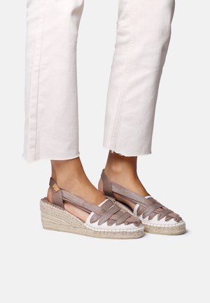 Espadrille wedges featuring a woven jute sole, metallic brown straps, open heel design, and white accents, set against light pants.