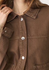 Giacca di denim marrone con colletto, bottoni a pressione argento e due tasche frontali con finitura testurizzata. Il tessuto ha una trama robusta.