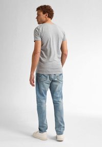 Tricou gri cu mânecă scurtă, blugi deschiși la culoare, uzati, și adidași albi; stând cu spatele întors, arătând un design și o croială casual simple.
