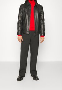 Veste en cuir noire avec fermeture éclair, associée à un col roulé rouge et un pantalon gris. La tenue comprend des bottes noires et un design épuré.