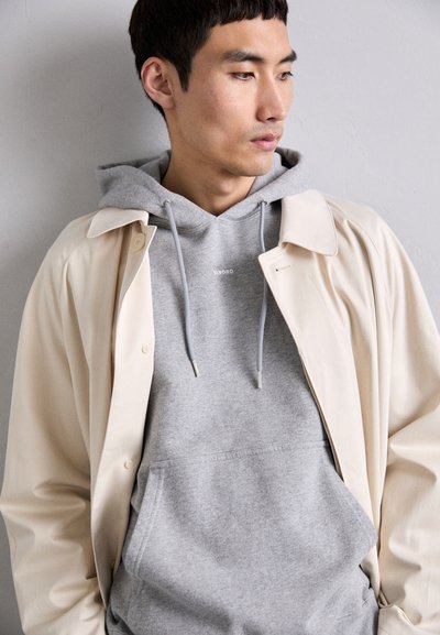 Couche extérieure beige à boutons sur un sweatshirt gris avec cordons de serrage, comportant une poche avant. Texture lisse pour les deux vêtements.