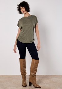 Camiseta texturizada de color verde oliva con mangas cortas, combinada con vaqueros skinny oscuros y botas altas de tacón color marrón claro. La modelo posa en interiores.