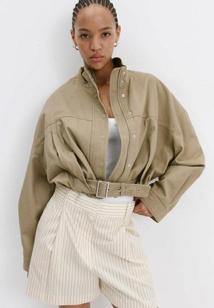 Mujer joven con pelo trenzado que lleva una chaqueta beige con cinturón, camiseta blanca y pantalones cortos de talle alto a rayas crema sobre fondo blanco.