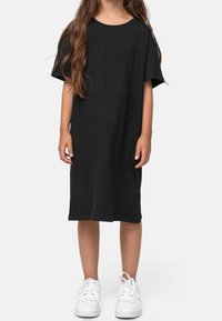 Enfant portant une robe noire unie à manches courtes et longueur genou, ainsi que des baskets blanches, debout devant un fond clair.