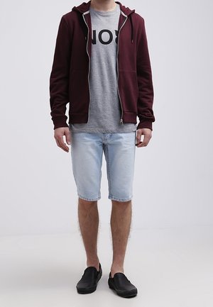 Sudadera burdeos con cremallera sobre una camiseta gris con letras negras grandes, pantalones cortos de mezclilla azul claro y zapatos negros sin cordones.