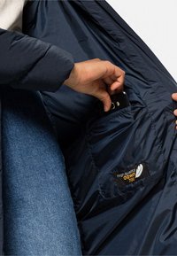 Dunkelblaue, gepolsterte Jacke von innen mit einer Tasche, die ein Smartphone hält, einem Etikett mit der Aufschrift "hochwertige Daune 700" und einer glatten Stoffstruktur.