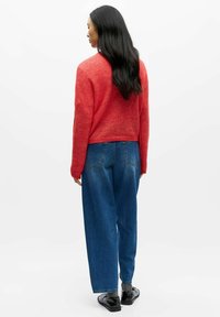 Sarkans vilnas maisījuma džemperis ar īsāku dizainu, kopā ar augsta vidukļa zilajām džinsu biksēm. Modelis valkā melnus apavus un ir ar gariem, taisniem matiem.