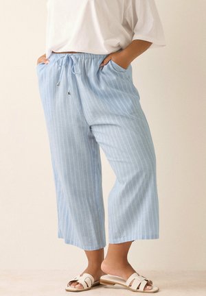 Persona con pantalones holgados de rayas azul claro con cordón y sandalias blancas, de pie con las manos en los bolsillos.