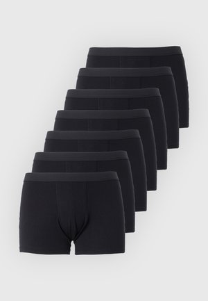 Štíhly mladý muž stojí bosý, v čiernych boxerkách na jednoduchom svetlom pozadí, pozerá sa do strany.