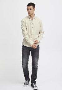 Chemise beige à manches longues avec des boutons, jean gris ajusté et baskets noires. La chemise présente une texture subtile et des boutons.