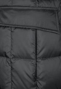 Zwarte pufferjack met gequiltte secties, een verticale rits en een klepzak op de borst, gemaakt van een zachte, lichte stof.