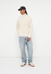 Maglione a collo alto lavorato a maglia crema abbinato a jeans baggy azzurri chiari e sneakers bianche. Un berretto nero completa il look con uno stile casual.