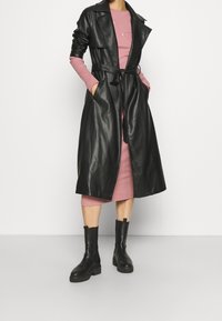 Svart läder trenchcoat med bälte i midjan, breda slag och sidofickor, lager på lager över en rosa kroppsnära klänning och svarta ankelskor.