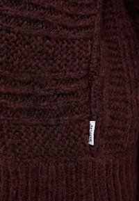 Burgundiško tono megztinis su tekstūriniais raštais ir keteriniais detalėmis. Maža balta etiketė su užrašu "Mustang" šone.